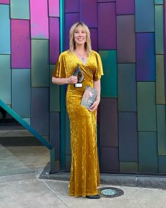 Trish Dool stands in front of a colorful block background. She is smiling at the camera, holding the Midwest Hairdresser award she won while wearing a golden yellow floor length velvet dress.
